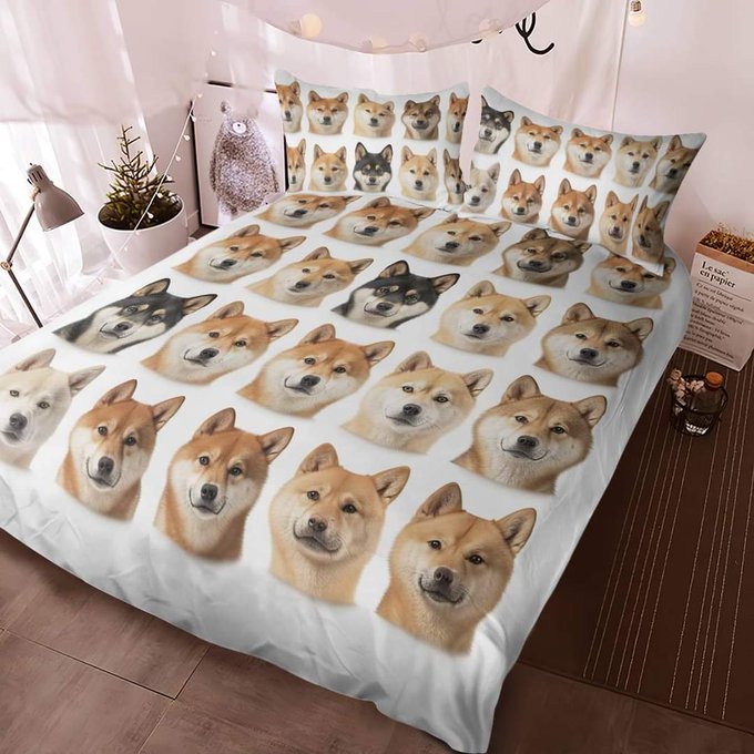 The image features a white bed with a comforter that has a unique design of various dog faces printed on it, including a brown dog face and other dogs' faces as well. The bed is set against a pink wall, which complements the white color of the bedspread. There are two pillows placed at the head of the bed, one with a brown dog face and another with a black dog face. A potted plant can be seen in the background on the left side of the image.