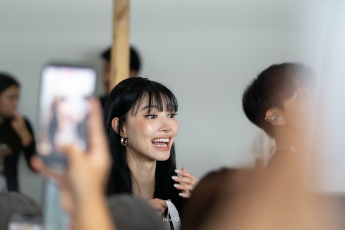 A candid shot of a smiling young woman with long dark hair and hoop earrings, surrounded by people and a phone camera capturing the moment, with the watermark 'isnk♡' at the bottom.