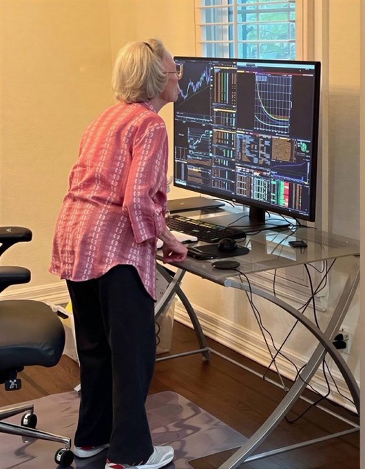 An elderly woman with short gray hair, wearing glasses and a pink patterned blouse, stands at a glass desk leaning forward to intently study multiple financial trading charts displayed on a large monitor.