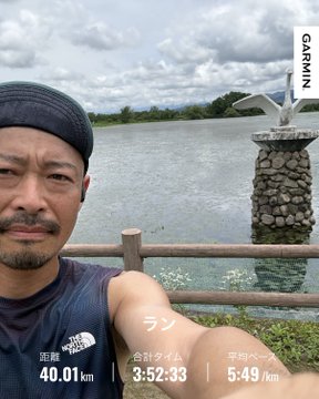 A man in a black beanie and The North Face shirt takes a selfie by a lakeside railing, with a seagull perched on a stone pillar in the background. The Garmin overlay shows "合計タイム 3:52:33", "平均ペース 5:49/km", and "距離 40.01 km", reflecting his long #STEPN run the day after drinking, which left him feeling dehydrated as noted in the post.
