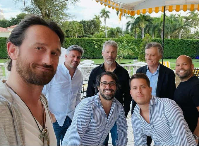 Seven men, including Michael Saylor and former Argentine President Mauricio Macri (flanked by bodyguards), pose together under a yellow-and-white striped canopy during a meeting arranged at The Bitcoin Conference in Miami.