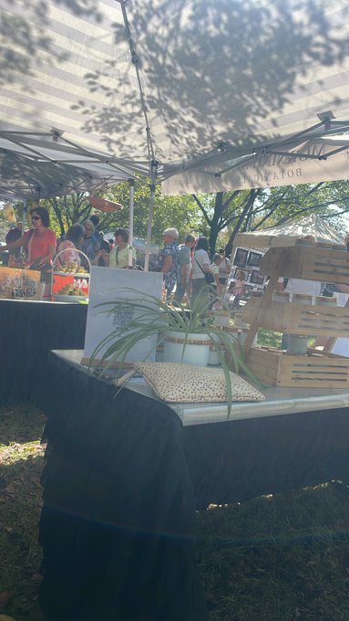 An outdoor market scene under a white tent canopy showing wooden crates, potted plants, and various shoppers browsing booths in a sunny setting.