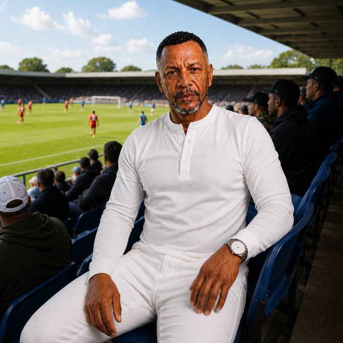 A man with a gray beard wearing a white long-sleeve henley shirt sits in stadium seats watching a soccer game in the background.