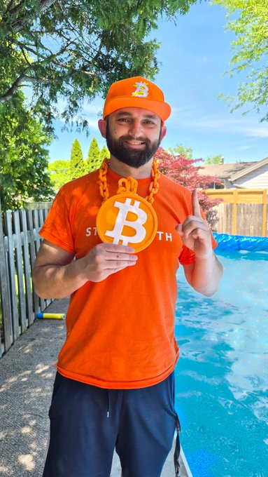 iced tea man holds a large orange "S" with a white "B" on it, standing by a pool and wearing an orange shirt and hat.