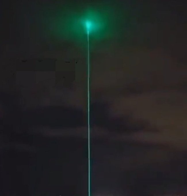A bright green laser beam shoots up into a dark, cloudy night sky, creating a glowing point at the top.