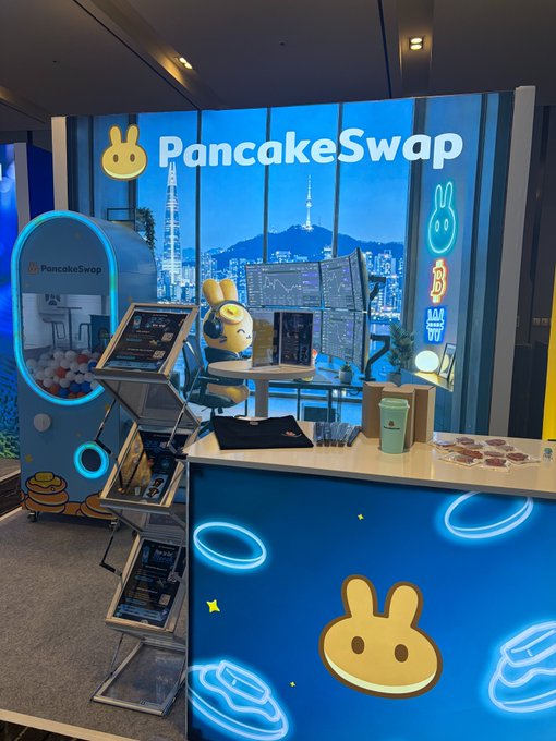 The PancakeSwap booth is set up at KBW (Korea Blockchain Week), featuring its iconic bunny logo, a blue themed backdrop with city skyline imagery, and a gacha machine for visitors to spin and win exclusive merch.