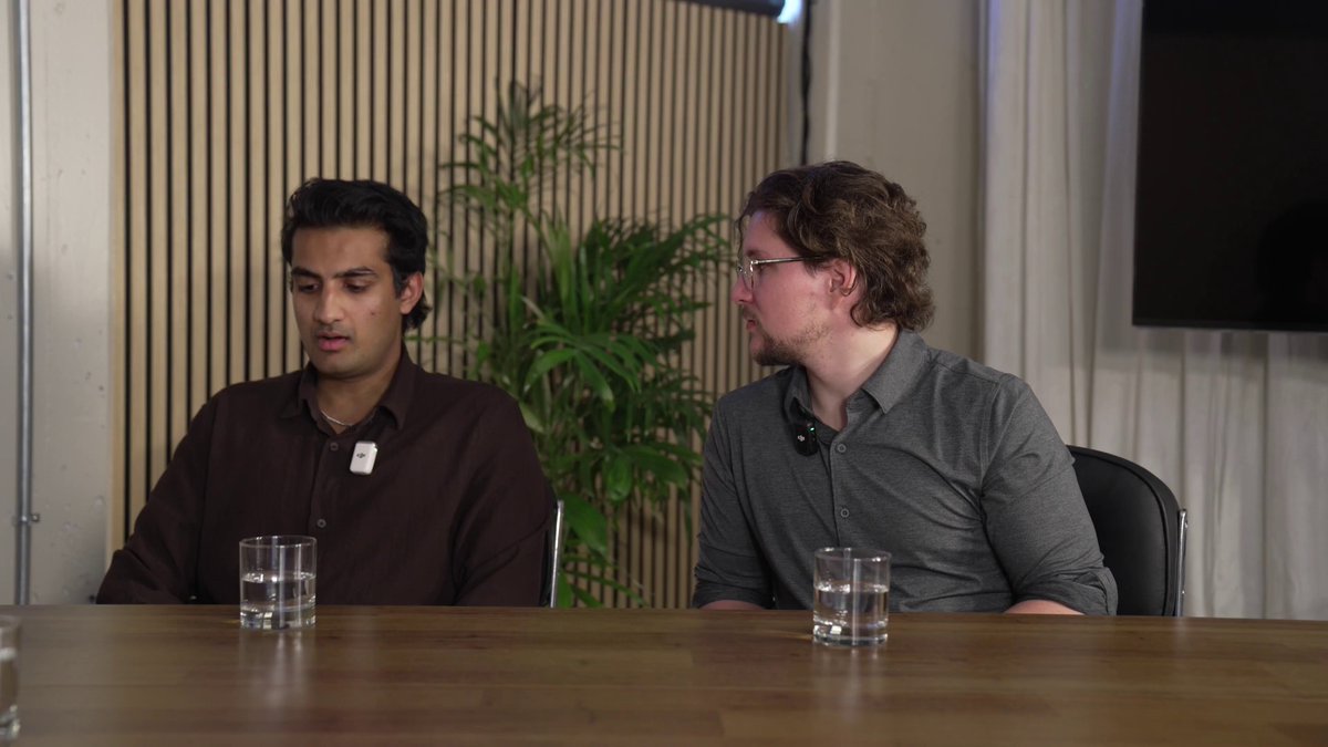 The image shows two men sitting at a table with glasses of water between them. They appear to be engaged in conversation, possibly discussing an interesting topic or sharing ideas. One man has his back turned towards the camera while the other faces the camera directly. The scene takes place in what appears to be a room with a TV and a potted plant nearby.
