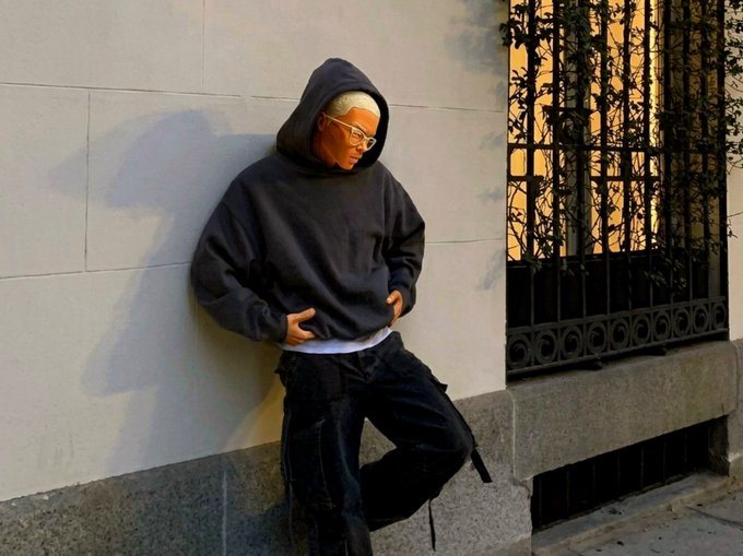 A young person with glasses and a beanie wearing an oversized dark hoodie leans against a light-colored wall on a street, posing with hands near their pockets.