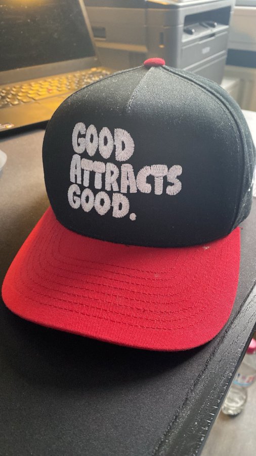A black and red baseball cap with embroidered text reading 'GOOD ATTRACTS GOOD.' sitting on a desk.