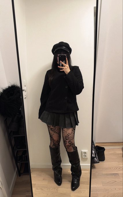 A person taking a mirror selfie wearing a black beret, black jacket, black pleated skirt, patterned tights, and tall black boots.
