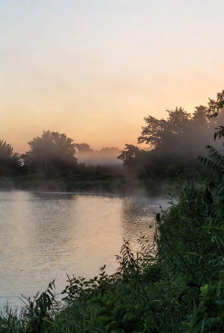 The image shows a serene river scene at sunrise, with fog rising over the water's surface. The sky above is painted in shades of orange, creating a beautiful backdrop for the tranquil setting. In the foreground, there are trees lining the riverbank, adding depth and texture to the landscape. The photo appears to be taken from a distance, capturing the entire scene without any specific focus on a particular subject or landmark.