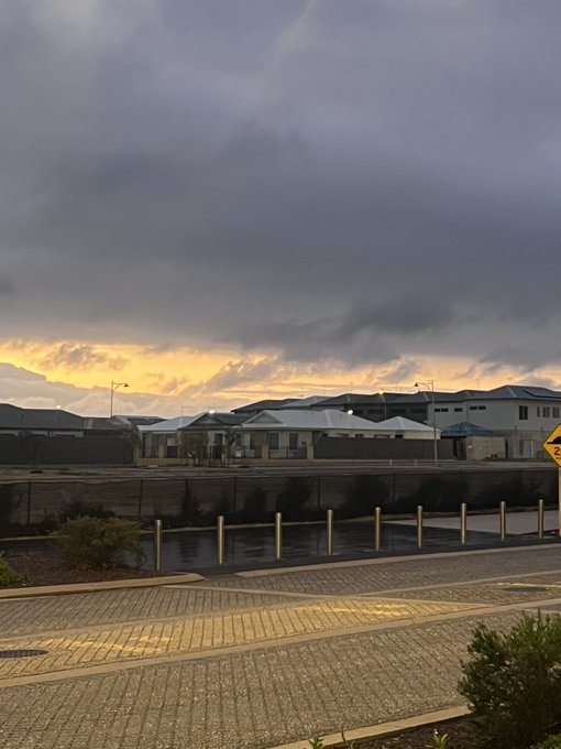 A scenic sunset view shows a cloudy sky with orange light near the horizon, overlooking buildings, a road, and a waterfront with railings.