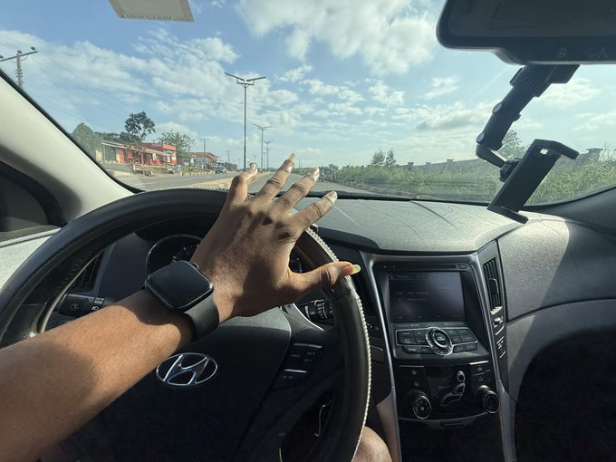A first-person view from inside a Hyundai vehicle, showing a hand resting on the steering wheel while driving along a road with solar streetlights under a partly cloudy sky.