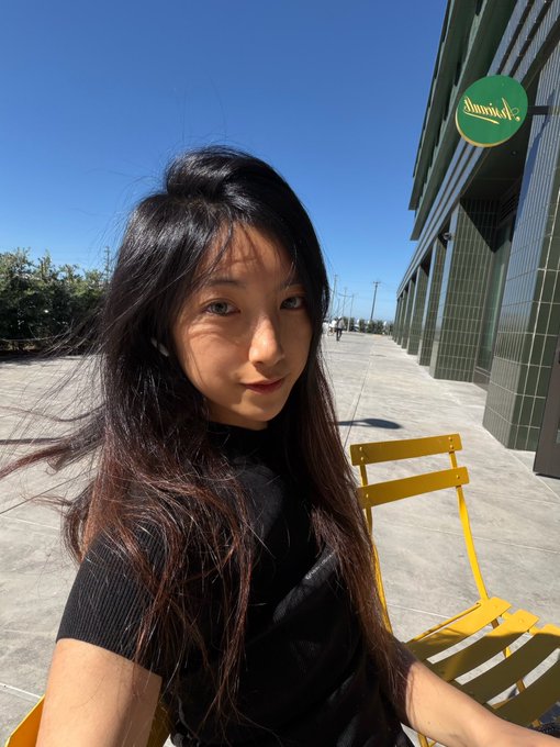 A selfie of a young woman with long dark hair sitting on a yellow chair outdoors on a sunny day, with a green building and a 'Lemonade' sign in the background.