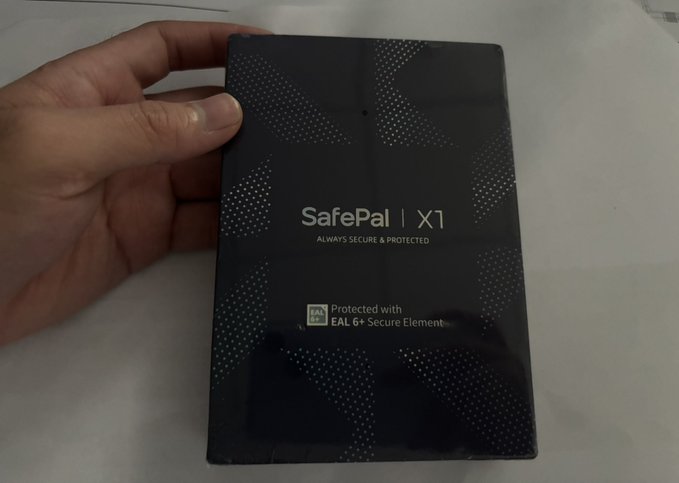 A hand holds a black "SafePal X1" hardware wallet package, featuring the tagline "ALWAYS SECURE & PROTECTED" and an "EAL 6+ Secure Element" label—which becomes ironic given the post's news that SafePal leaked data of nearly 40,000 customers.