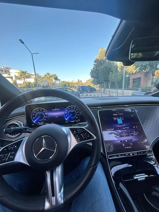 Interior view of a Mercedes-Benz vehicle showing the steering wheel, digital dashboard, and central infotainment screen with a parking lot and palm trees visible through the windshield.