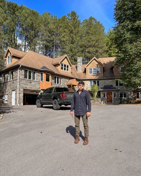 A man stands proudly in front of a sprawling, off-grid stone mansion surrounded by forested acreage, with a pickup truck parked behind him—visually embodying the post's themes of estate acquisition, seclusion, and self-made wealth.