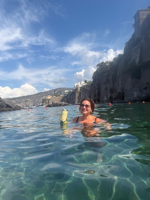 A woman with sunglasses smiles while relaxing in clear turquoise water along a rocky coastline, holding up a yellow beverage can, with colorful buildings perched on the cliffs in the background.