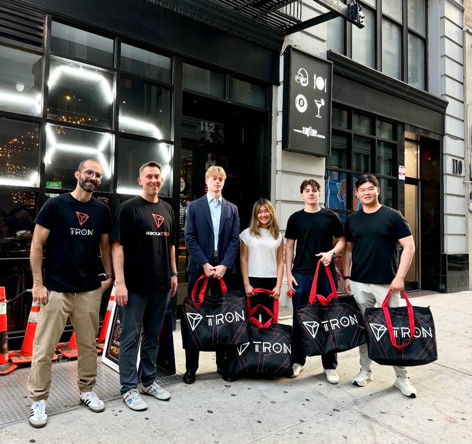 The image features a group of six people standing outside a building, posing for a photo with their bags. The bags are black and white, which matches the color scheme of the post that says "TRON Academy". The text on the post is in Chinese, but the rest of it is not visible in this particular image. The people in the group appear to be students or participants at the TRON Academy, as they have their bags with them.