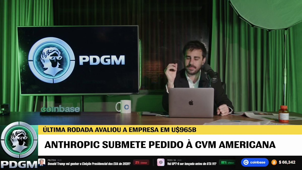 The image features a man sitting at a desk with his laptop open, possibly working or browsing the internet. The background of the image shows a green curtain, which could be part of an office setting. There are also two TVs visible in the scene - one is located behind the man and another on the left side of the image. A cup can be seen placed near the center of the desk.
