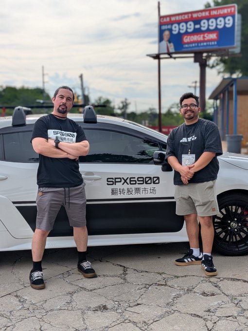 Two men pose in front of a white car labeled "SPX6900 翻转股票市场" (Flipping the Stock Market), representing the SPX6900 ticker that's bringing the community together as a meme stock movement. Behind them, a billboard advertises "George Sink Injury Lawyers" with the phone number 999-9999, providing a coincidental backdrop to this crypto/stock culture meetup.
