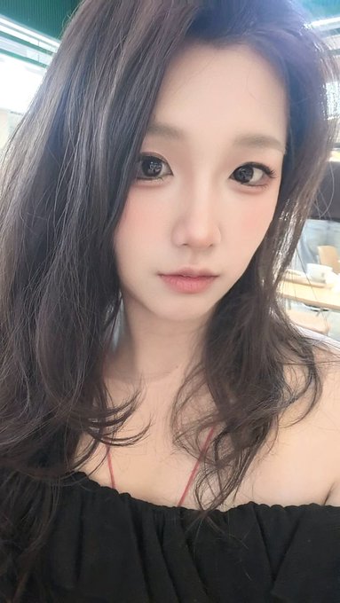 A close-up selfie of a young woman with long dark hair wearing a black off-shoulder top.