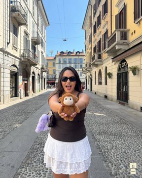 A woman playfully holds up a "MonkeBubus" plush toy in a charming Milan street, mirroring the post's narrative of these toys being spotted and "captured" globally. She echoes the idea of these plushies taking over, as one is already attached to her handbag.