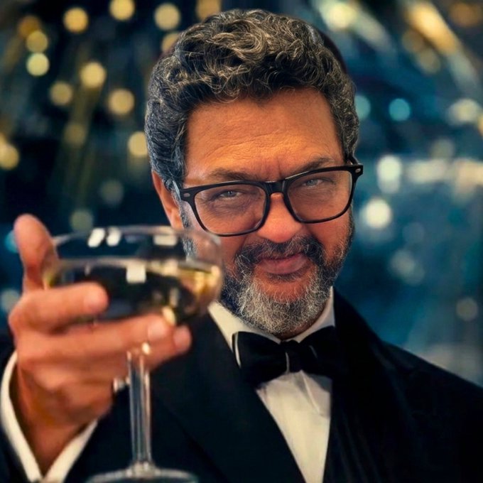 A portrait of a smiling, bearded man wearing glasses and a black tuxedo with a bow tie, holding up a glass of white wine against a bokeh background.