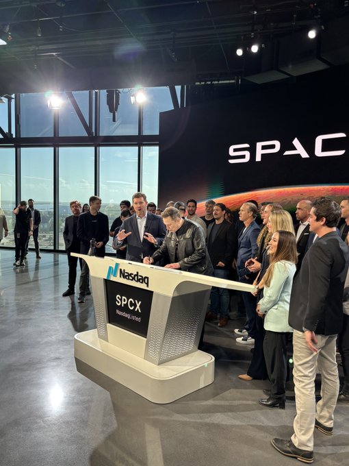 The image shows a group of people gathered around a table, with one man standing at the head of the table and others sitting or standing behind him. The table has a sign that reads "SPACX" on it. A person is giving a speech in front of this gathering, which suggests they are addressing an important topic related to the company's future plans or announcements.