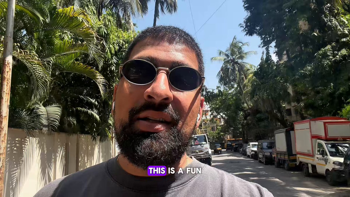 The image shows a bearded man wearing sunglasses, likely taking a photo of himself with his surroundings, including several parked vehicles such as trucks and cars around him. The surrounding area appears to be a street or parking lot with palm trees visible behind a fence in the distance.