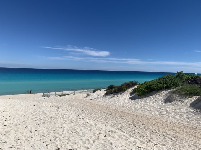 Tropical beach scene featuring white sand dunes, turquoise water, and a clear blue sky with wispy clouds.