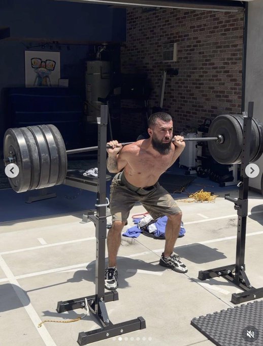 A shirtless, bearded man performs a heavy barbell squat in an outdoor gym setting, with multiple weight plates loaded onto the bar.
