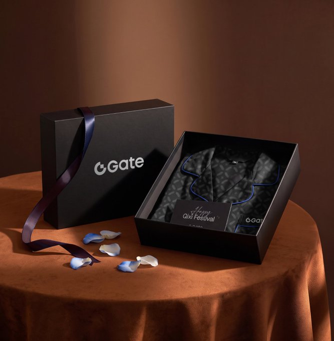 Elegant product shot of a black Gate gift box with ribbon alongside an open box revealing a patterned silk garment and a 'Happy QIXI Festival' card, displayed on a brown surface with scattered flower petals.