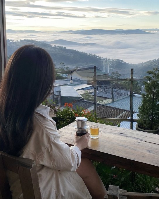 A woman with long dark hair sits at a wooden table with a coffee dripper and glass, overlooking a misty mountain landscape at sunrise.