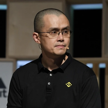 Portrait of Changpeng Zhao (CZ), the Binance CEO, wearing a black polo shirt with the Binance logo and glasses, speaking at an event.