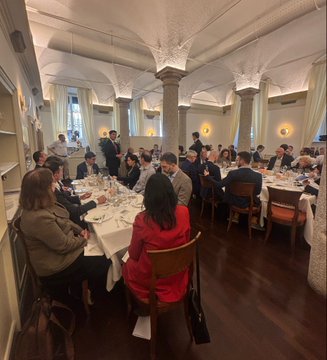 A group of people are seated at round tables in an elegant dining room with ornate columns, attending what appears to be a formal gathering or meal.