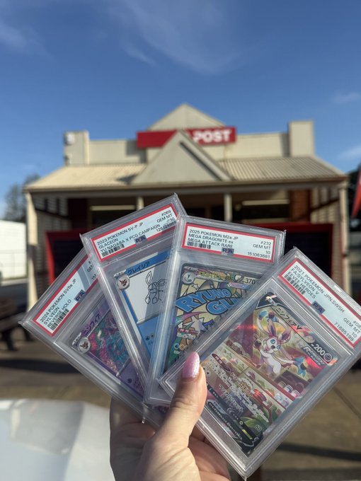 Hand holding several PSA-graded Pokemon trading cards in protective slabs, including a Sylveon ex card, photographed in front of a building marked 'POST'.