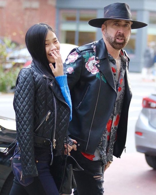 A woman in a black quilted leather jacket laughs while holding hands with a bearded man wearing a wide-brimmed hat and a floral-painted leather jacket, standing on a city street. The image humorously illustrates the caption's vision of a 2045 future where crypto fortunes funded a life of idle luxury in Thailand, with the couple's laughter mocking the absurdity of pretending their life's work amounted to anything more than a "middling outcome."