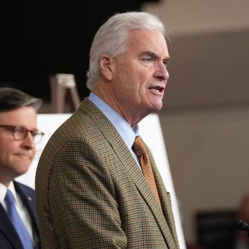 Rep. Tom Emmer, wearing a checkered tan blazer and orange tie, speaks at a podium during a public appearance, with another bespectacled official seated behind him. The image accompanies news of his comments to Fox Business about the CLARITY Act's delayed cloture vote following the midterms.