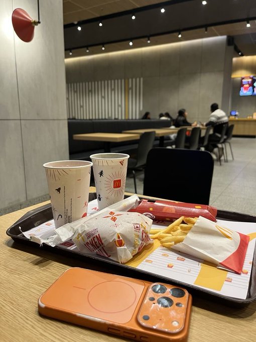 A McDonald's breakfast meal sits on a tray, featuring two paper cups with the iconic "M" logo, a wrapped burger, fries, and an orange phone case resting on the table. The "Good morning CT" caption pairs perfectly with the scene of a quick breakfast stop in the modern fast-food restaurant.
