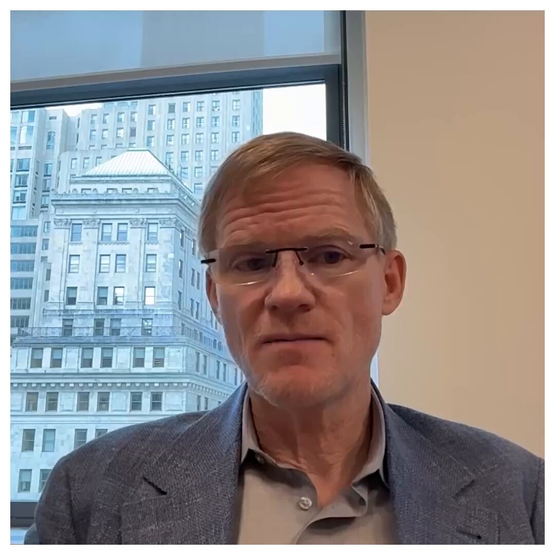 The image features an older man wearing glasses sitting at his desk with a window behind him that overlooks a city skyline. The man appears to be dressed professionally, possibly in a suit or blazer, which suggests he might be a business professional. He is looking directly into the camera and seems to be engaged in conversation or contemplating something. The presence of the city skyline in the background indicates that his workplace could be located in an urban setting with tall buildings nearby.