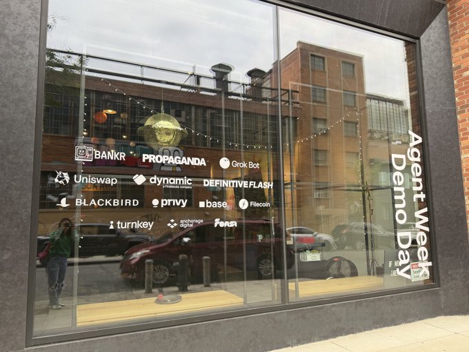 The storefront window displays logos of various crypto and fintech companies (including Uniswap, Bankr, Propaganda, Grok Bot, Dynamic, Definitive Flash, Blackbird, Privy, Base, Filecoin, Turnkey, Anchorage Digital, and ENS) alongside bold text reading "Agent Week Demo Day," promoting a startup showcase event.