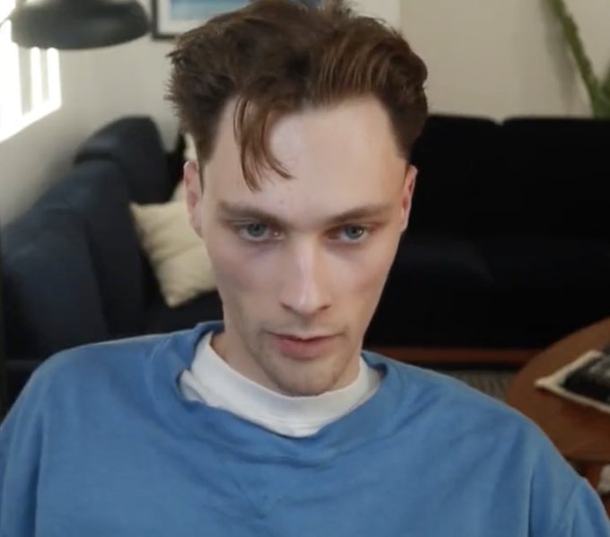 A young man with tousled brown hair and blue eyes stares directly at the camera with a serious expression, wearing a blue sweatshirt over a white tee in a softly lit living room. The accompanying post references "Subway Ticker font" and "@cryptopunks brand guidelines, 2026," suggesting the image may serve as a visual reference or mood board for branding work, though no text or branding elements are visible in the photo itself.