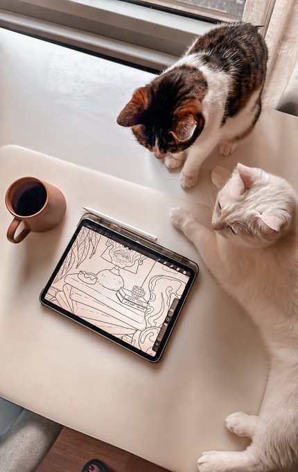 Two cats sit on a white table looking at a tablet displaying a digital coloring page, with a white stylus and a mug of coffee beside it.