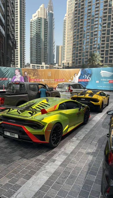 A bright green Lamborghini with "90955" on its plate is parked along a Dubai Marina street, with the caption's wordplay on "GM" playfully echoed by the "26" visible on the Gulf News billboard behind it. The luxury supercar scene captures Dubai's opulence while tying into the post's greeting pun.
