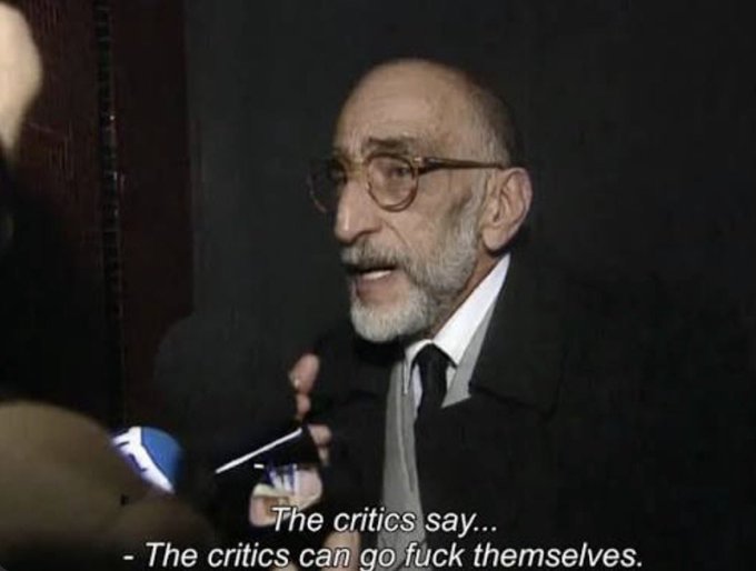 An elderly man with a white beard and glasses speaks defiantly into a microphone, with subtitles reading "The critics say... - The critics can go fuck themselves." The image serves as a humorous visual metaphor for the post's message about confidently dismissing critics and bots who spread misinformation about Azuki releases.