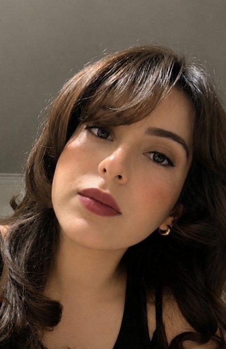 A close-up selfie of a young woman with long brown hair, blue eyes, mauve lipstick, and small hoop earrings, looking at the camera with a neutral expression.