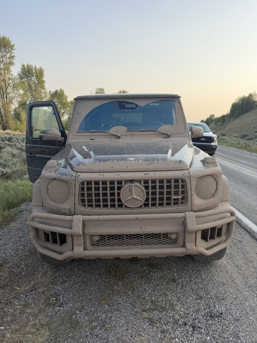 A Mercedes-Benz G-Wagon is covered in a thick layer of dust from off-road driving, with the driver's door open and another SUV visible in the background. The caption "Got a lil dirty" humorously understates the heavy dust coating that has nearly obscured the iconic Mercedes-Benz emblem on the grille.