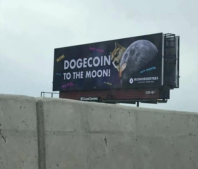 A billboard sits atop a concrete barrier, prominently displaying "DOGECOIN TO THE MOON!" in bold white letters against a starry space backdrop, accompanied by the Shiba Inu dog mascot reaching toward a detailed image of the moon, while text like "wow," "such crypto," and "much mooning" adds playful cryptocurrency slang, and the bottom corner credits "RUSHORDERTEES CUSTOM APPAREL" with the number "010-181," creatively illustrating the viral "Dogecoin to the moon" meme that references the cryptocurrency's aspirational price surge.