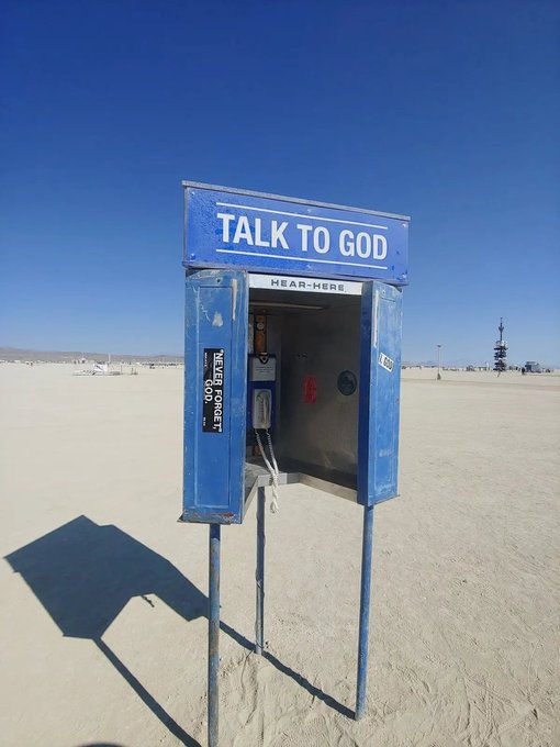 A blue 'TALK TO GOD' payphone booth stands in the middle of a vast, flat desert landscape under a clear blue sky.